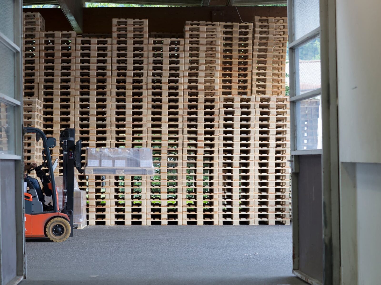 Forklift carrying a wrapped load beside stacks of timber pallets.